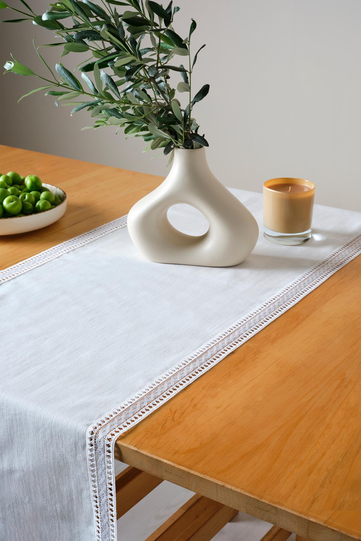 White lace table runner on a rectangular wooden table, featuring a modern vase, green fruits, and candle decor.